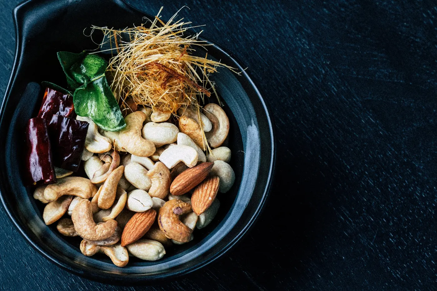 A dark bowl of mixed nuts and seeds with a dried chile