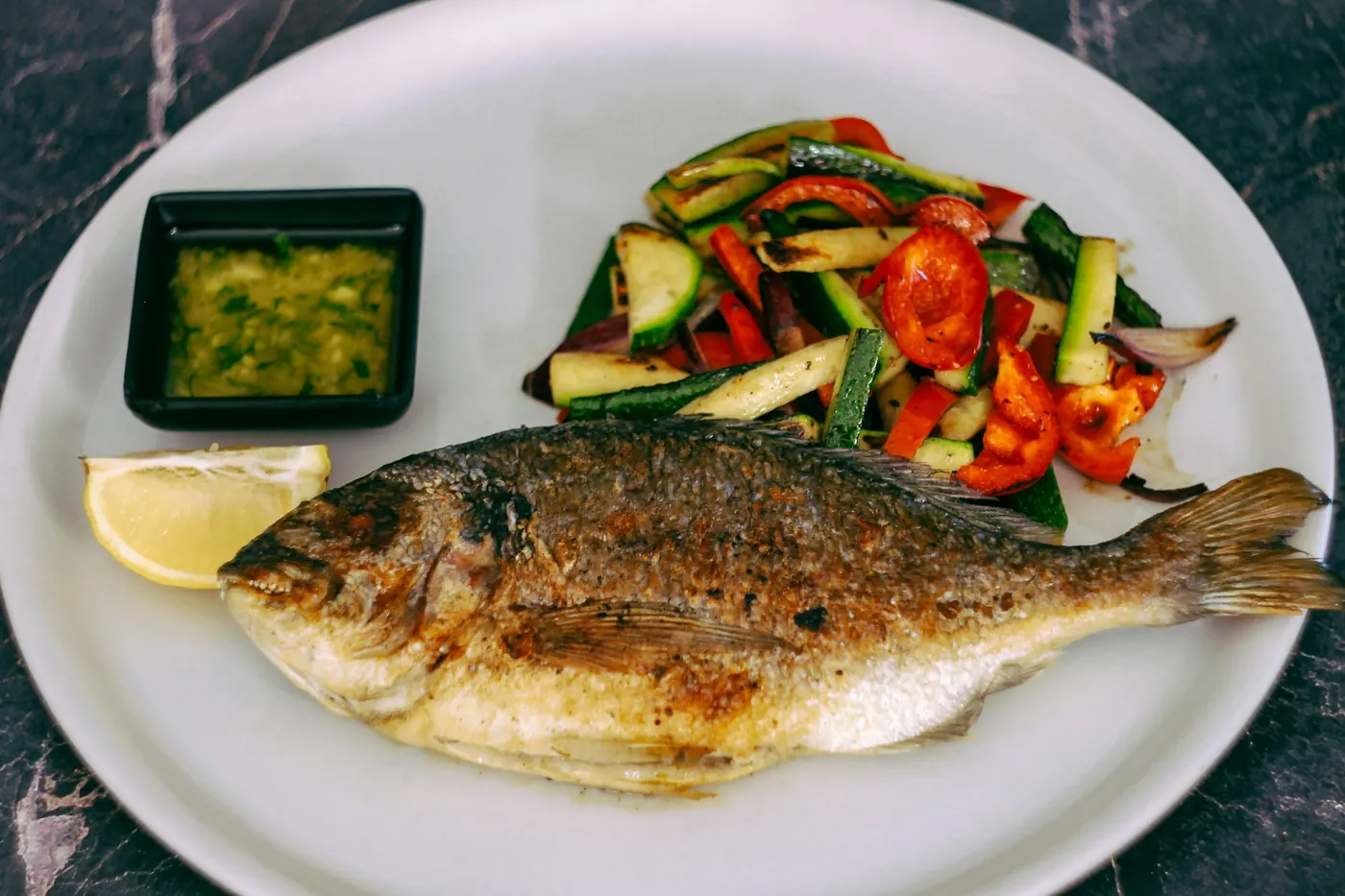 A grilled whole fish plated with vegetables and a green sauce
