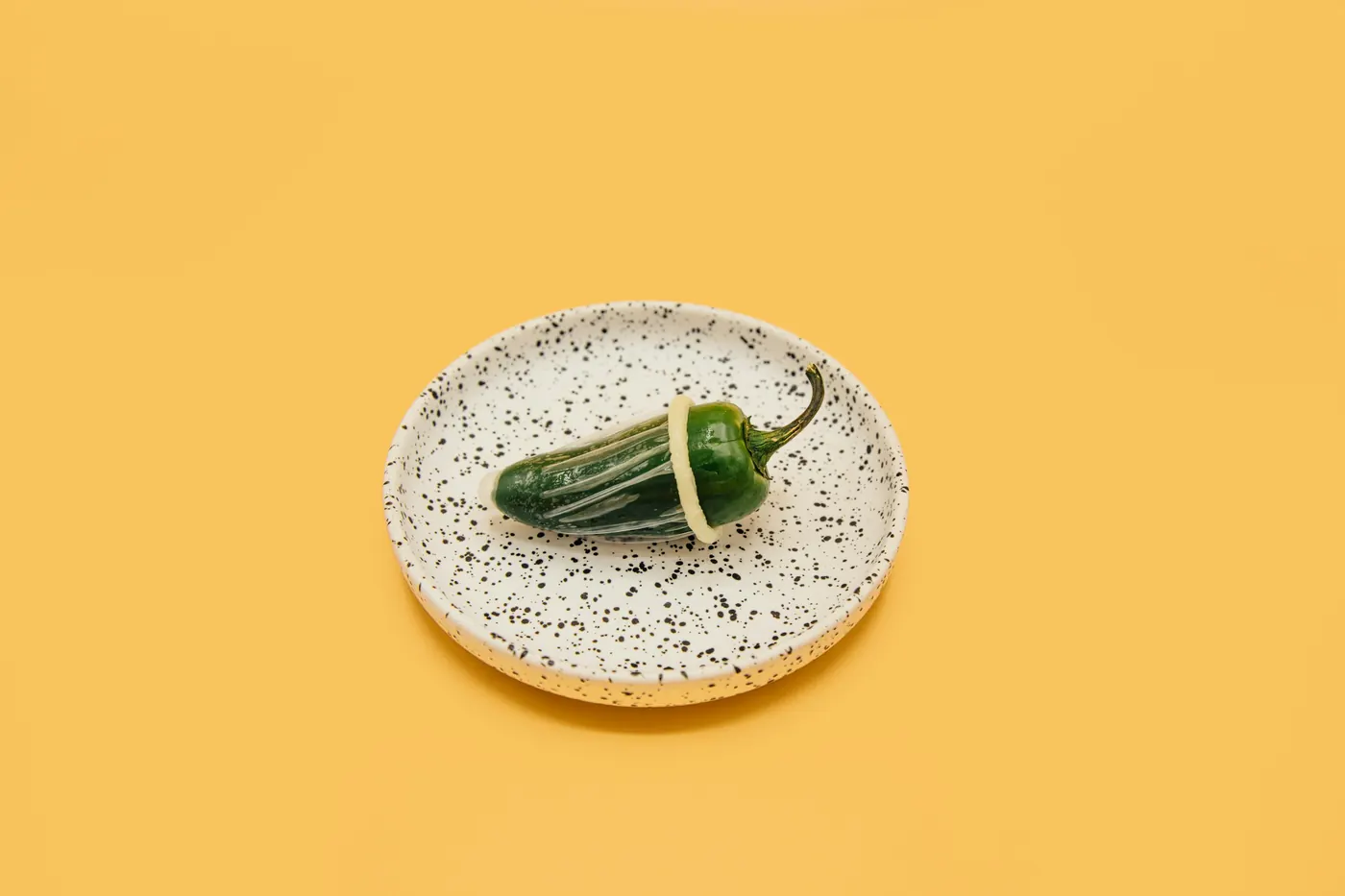 A single green chile pepper on a speckled plate against a yellow background