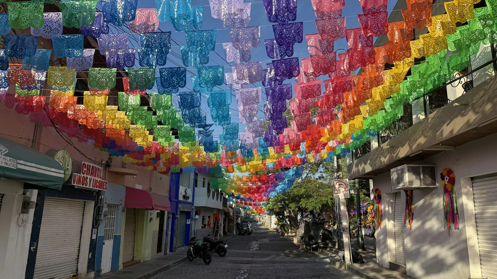 A narrow street covered overhead by rows of rainbow papel picado banners