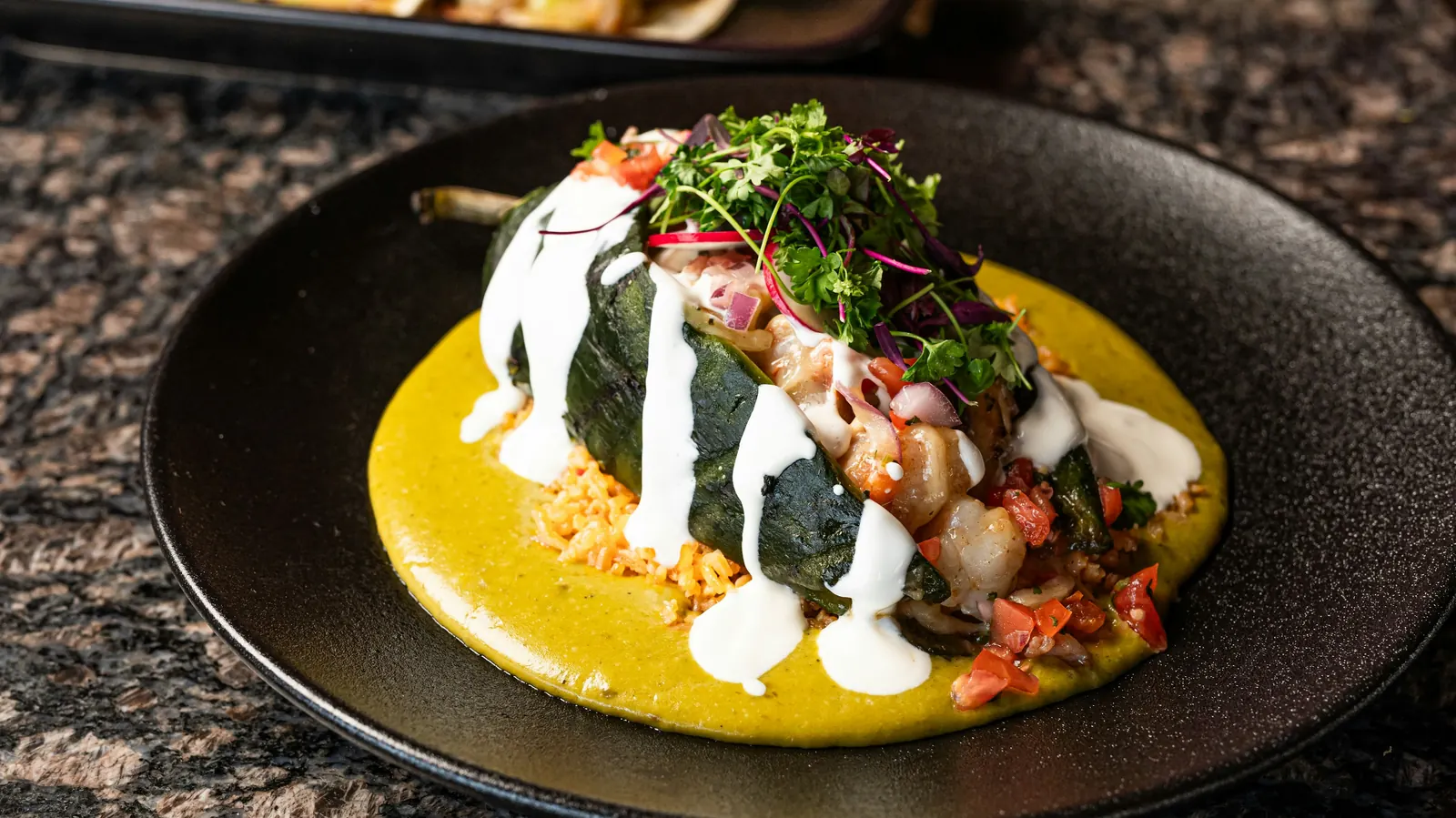 A whole stuffed green chile on a dark plate under a pale sauce with cream drizzled over
