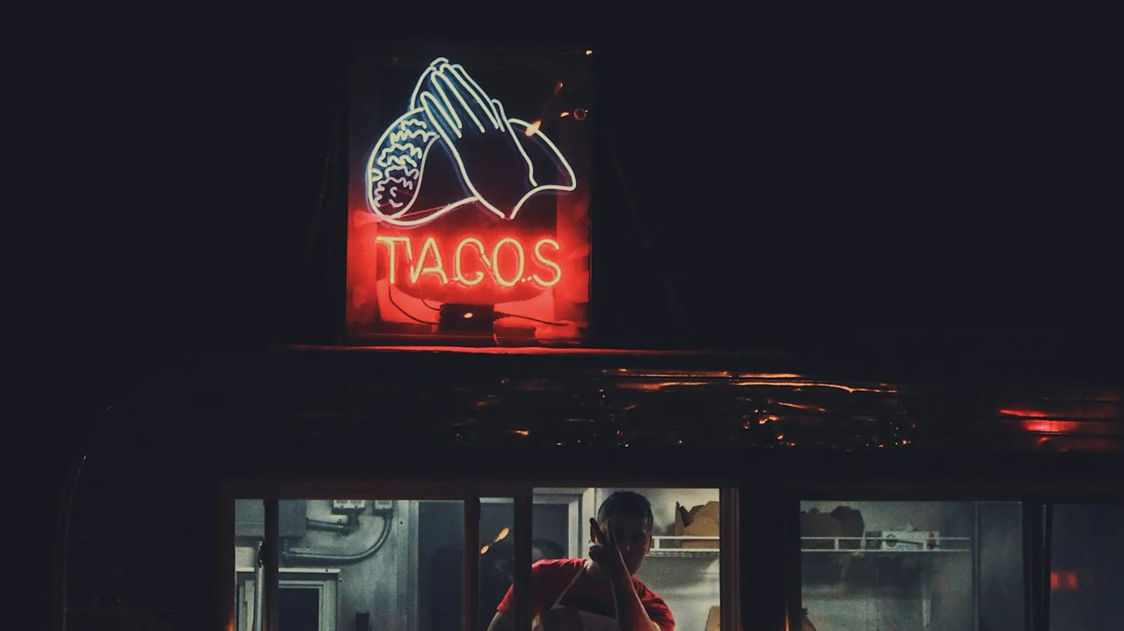 A lit sign reading tacos above a small building at night