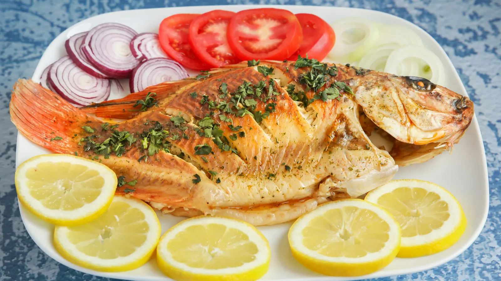 A whole cooked fish plated with lemon slices, tomato and onion