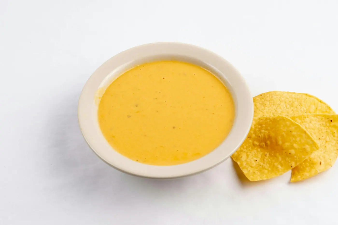 A white bowl of smooth yellow chile con queso with tortilla chips