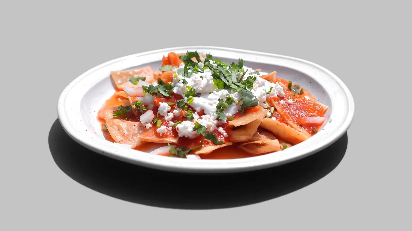 Tortilla pieces in red sauce on a white plate, topped with cream and crumbled cheese