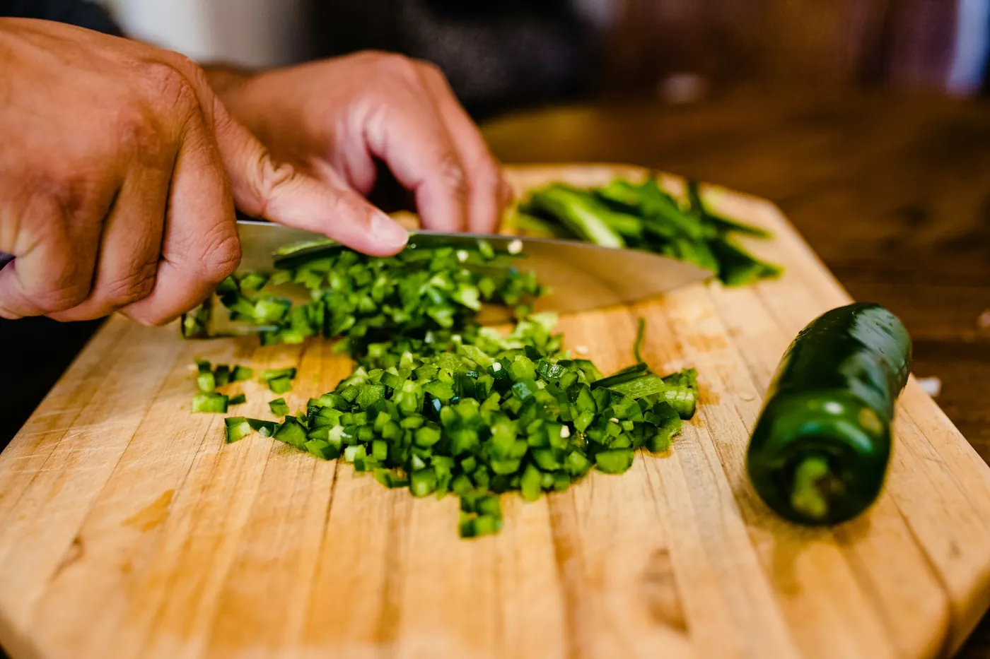 Hands finely chopping green chile on a wooden board beside a whole one