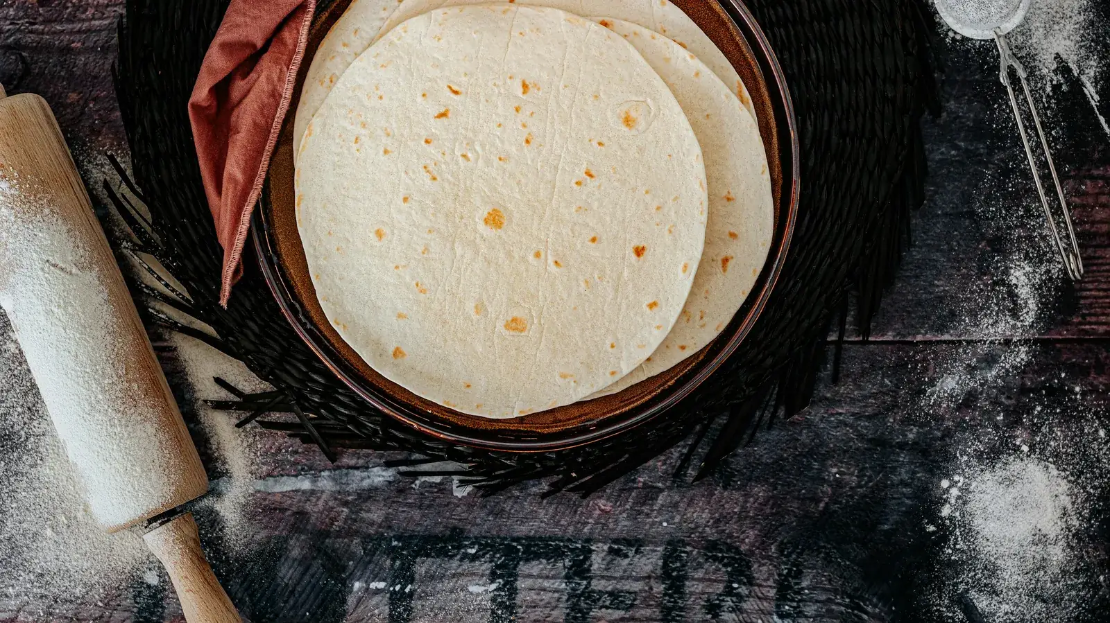 A stack of soft flour tortillas in a wooden bowl on a dark surface