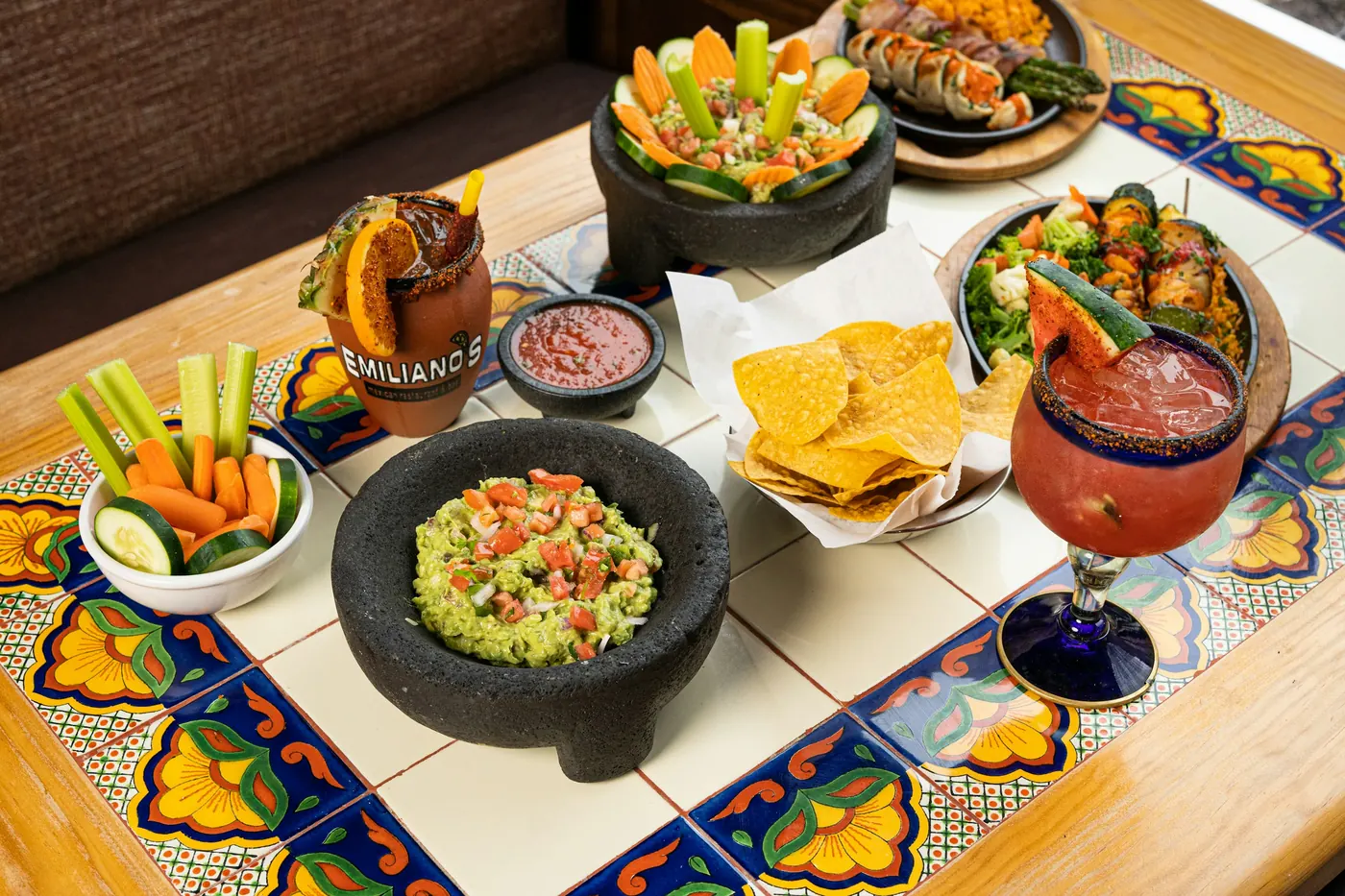A stone molcajete of guacamole with tortilla chips and drinks on a tiled table