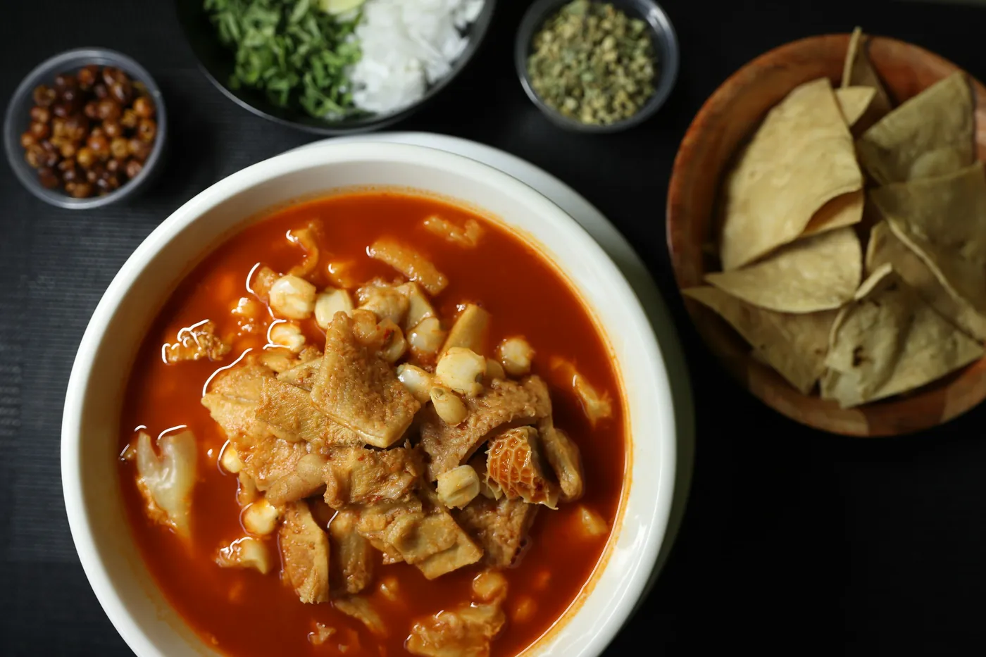 A bowl of red menudo soup containing pieces of tripe, with bay leaves alongside