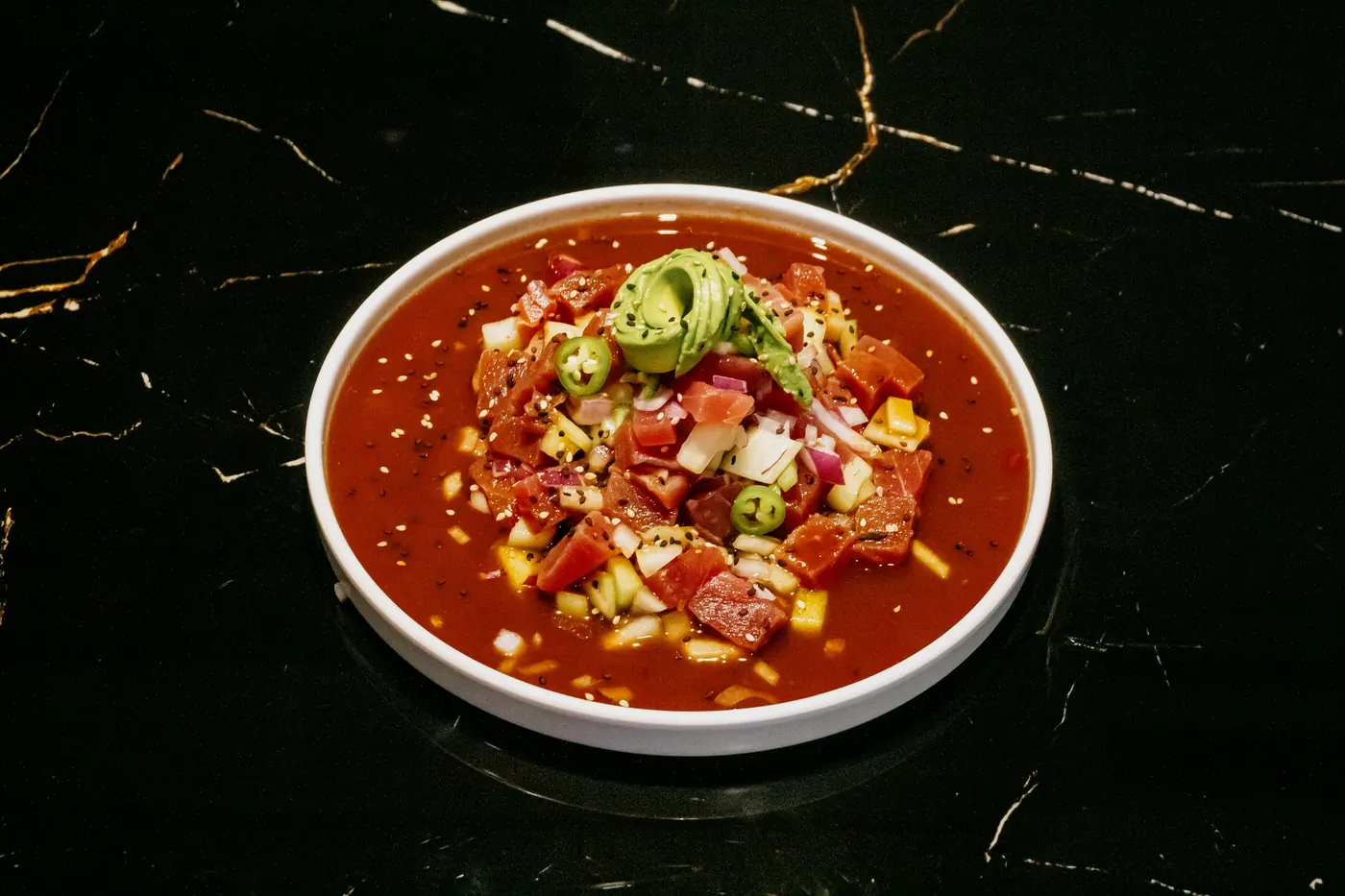 A red soup bowl topped with avocado and corn on a dark surface