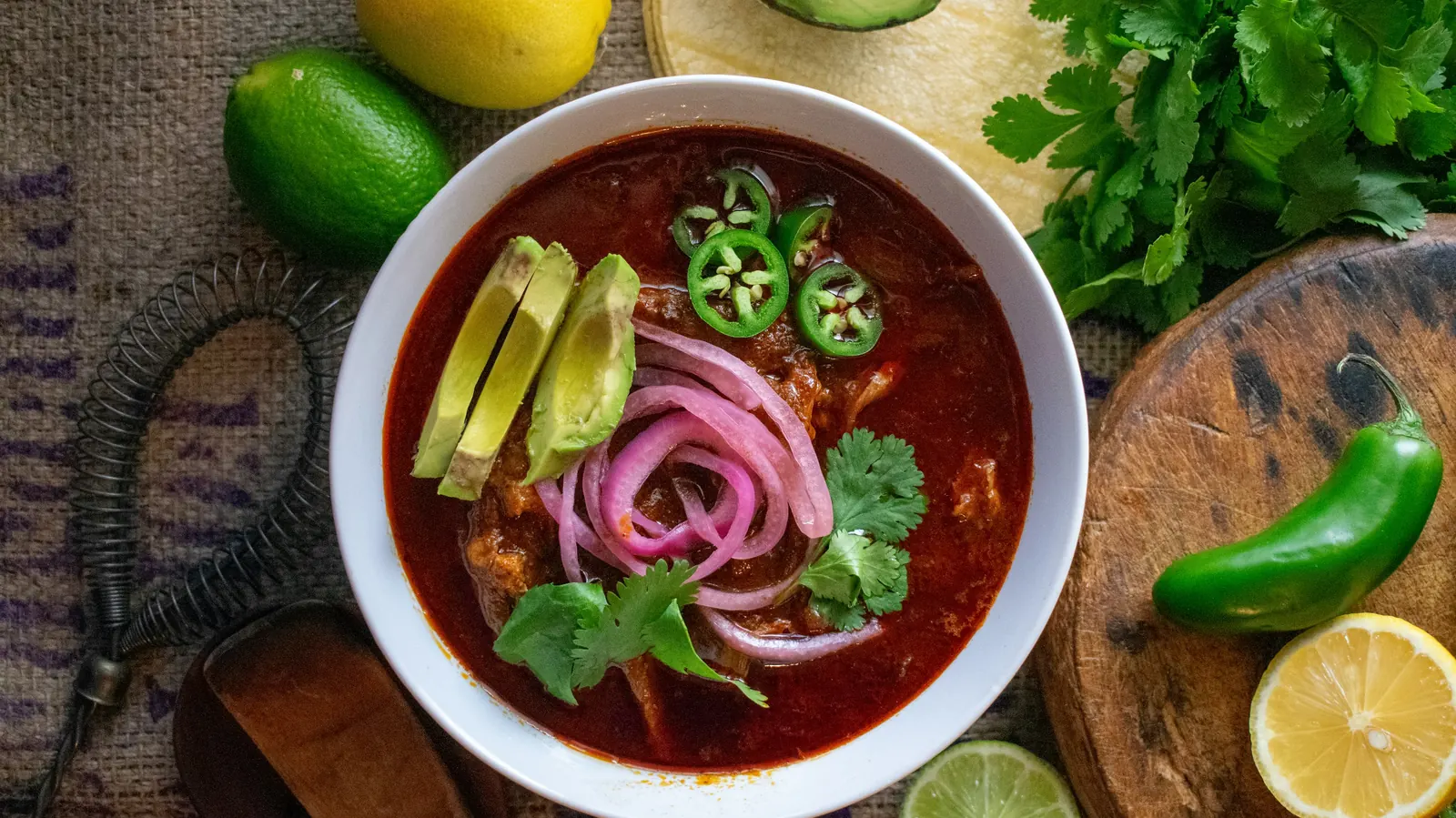A bowl of red Mexican soup topped with pickled onion, avocado and jalapeno