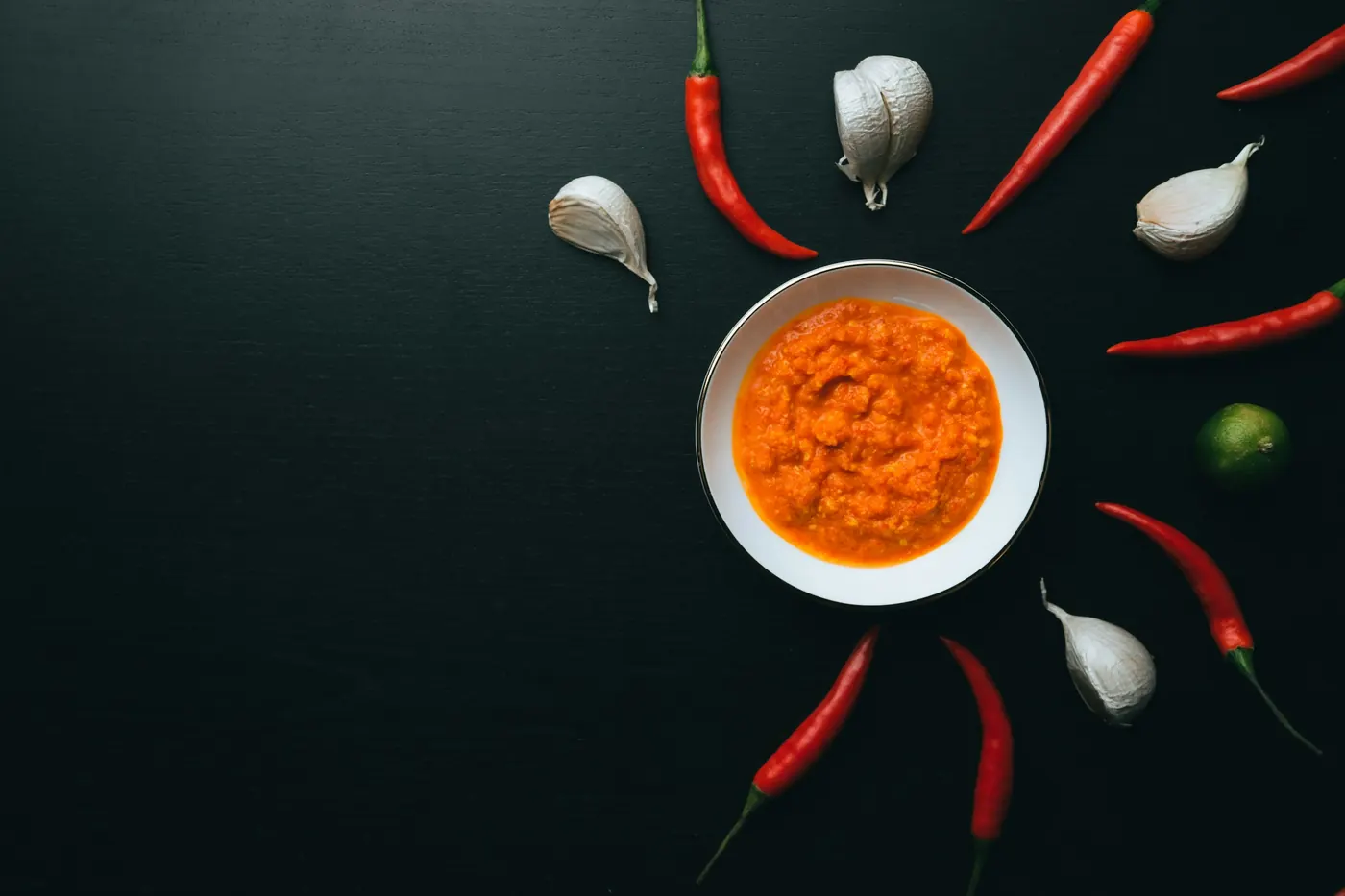 A bowl of red-orange sauce surrounded by dried chiles and garlic cloves