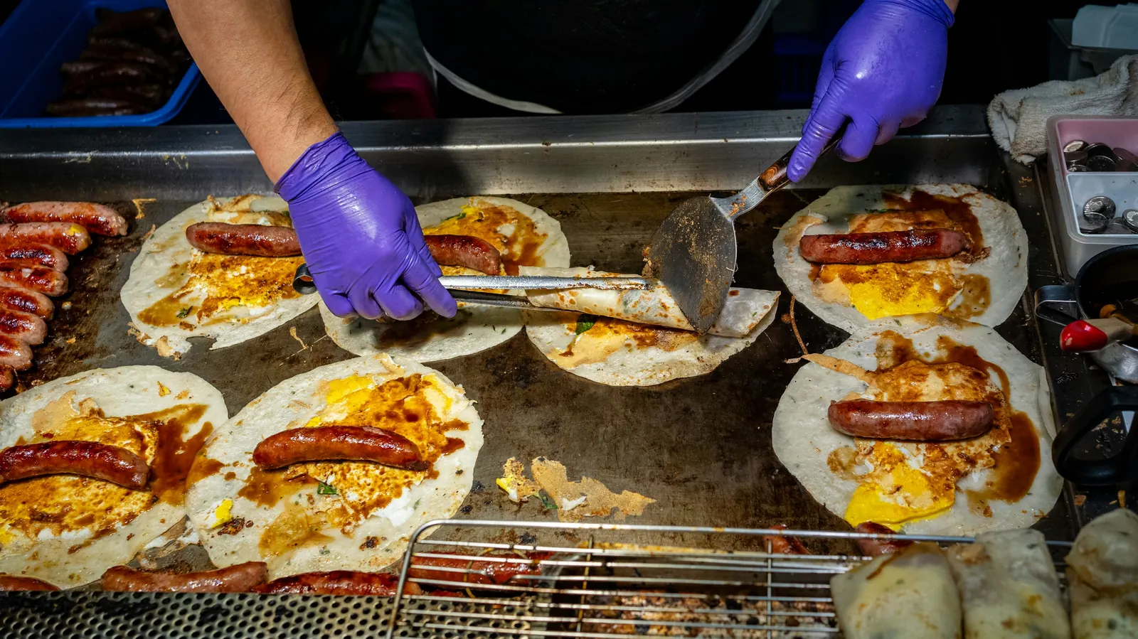 Gloved hands cooking eggs, sausage and tortillas together on a flat griddle