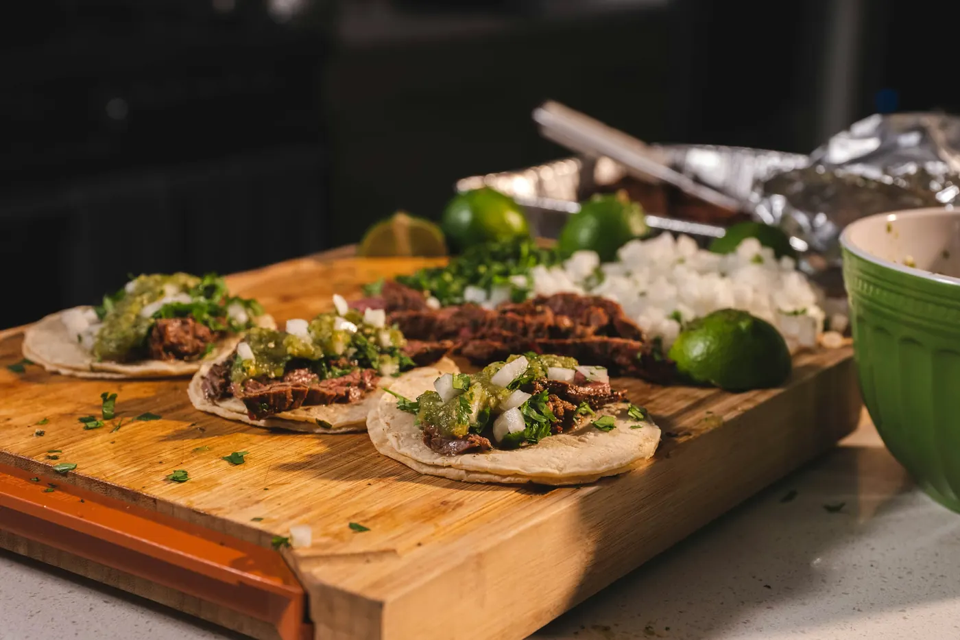 Small tacos lined up on a wooden board beside a sheet of foil
