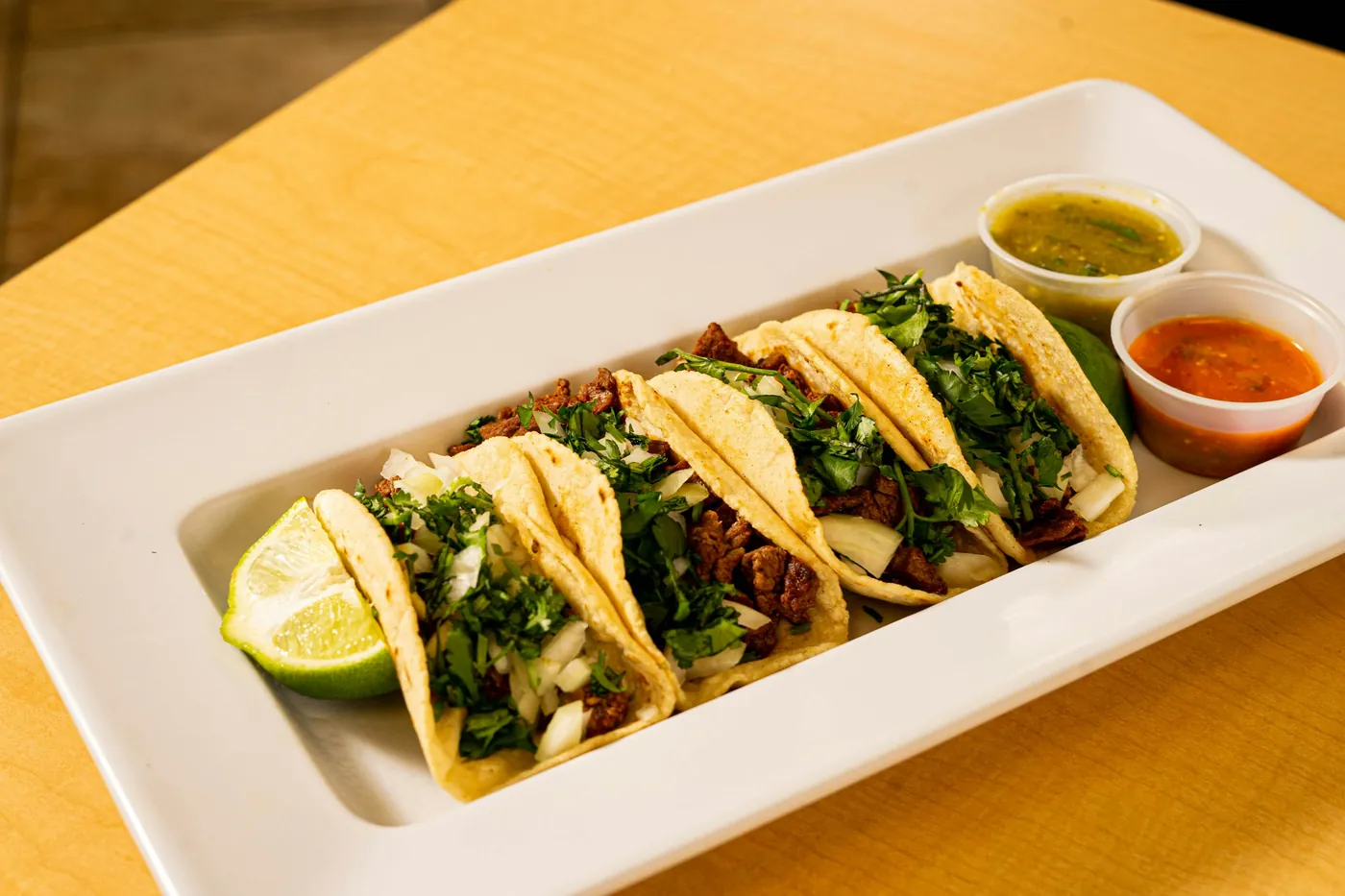 A row of tacos served on a long white platter with small bowls of salsa