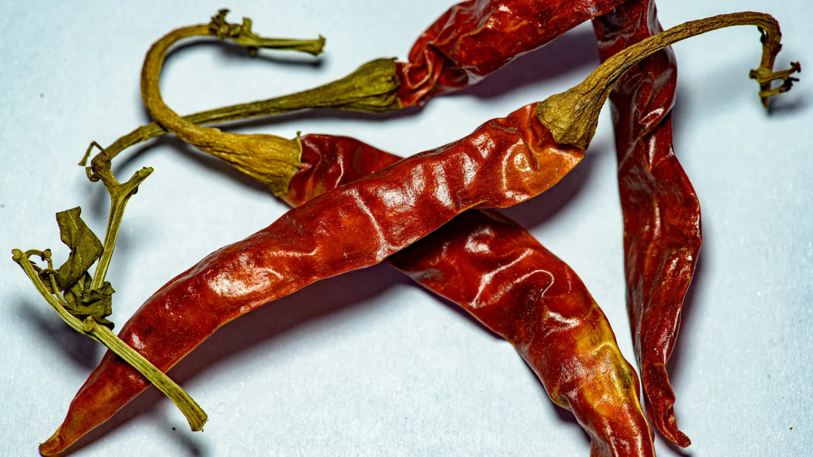Three long smooth dark red dried chiles with curled stems on a pale surface