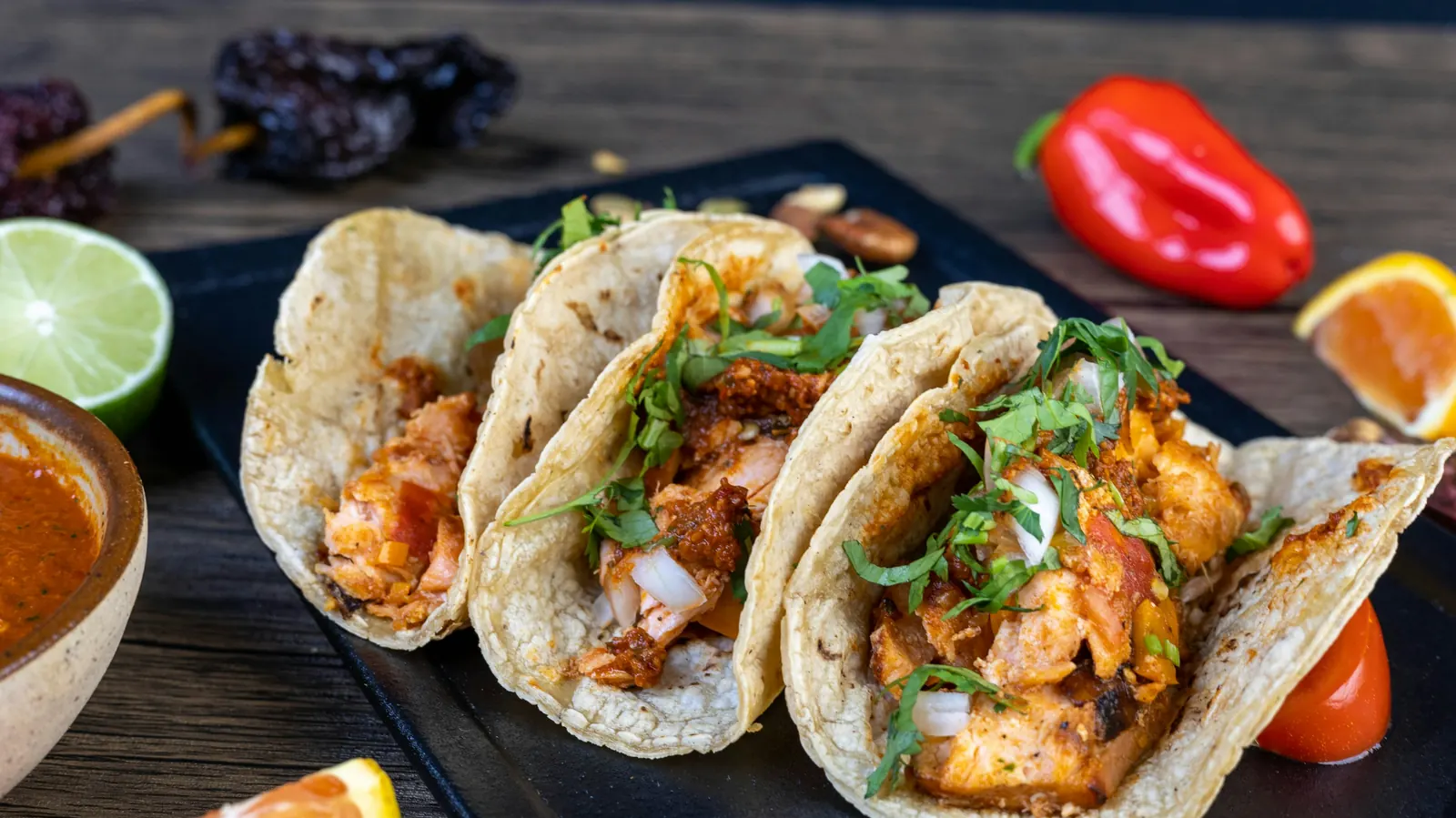 Three seafood tacos on a dark board with lime wedges and chiles
