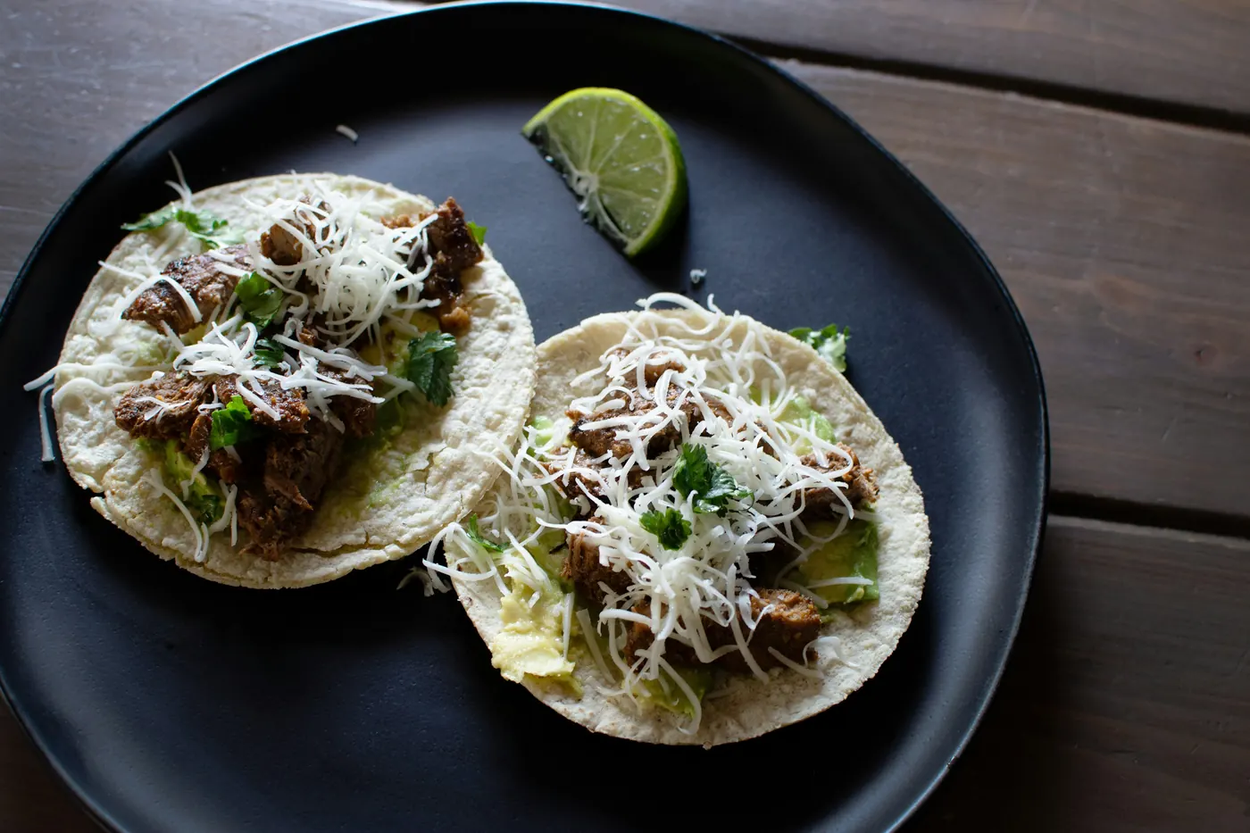 Two soft tacos with shredded meat and lime on a black plate